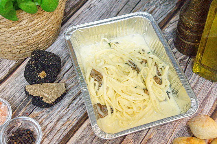Ein Folienbehälter mit cremiger Pasta und Trüffelscheiben steht auf einer Holzplatte, umgeben von einem Trüffel, Olivenöl, Salz, Pfefferkörnern, Brot und einer Topfpflanze.