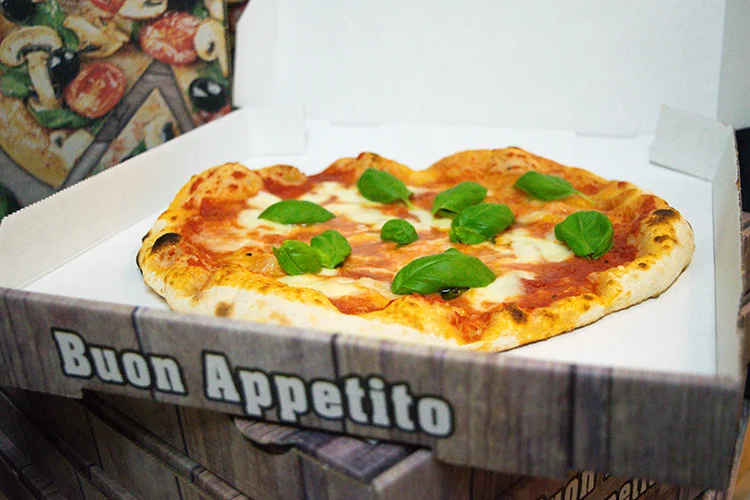 Eine Pizza mit geschmolzenem Käse, Tomatensauce und frischen Basilikumblättern liegt in einem offenen Pizzakarton mit der Aufschrift "Buon Appetito". Die Kruste ist golden und leicht verkohlt. Im Hintergrund ist eine andere Pizza zu erkennen.