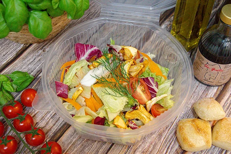 In einem durchsichtigen Plastikbehälter befindet sich ein bunter Salat mit Kopfsalat, Tomaten, Karotten und Kräutern. Um ihn herum stehen Kirschtomaten, Basilikum, Brötchen, eine Flasche Olivenöl und eine Flasche Balsamico-Essig auf einer Holzfläche.
