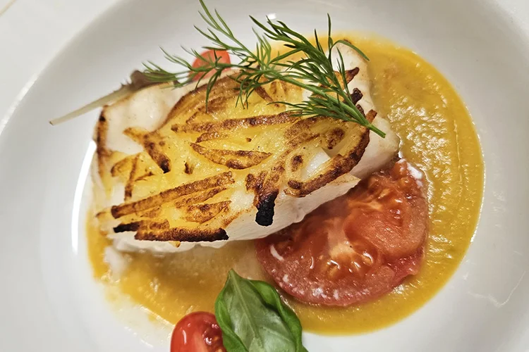 Ein Tellergericht mit gegrilltem Weißfischfilet auf gelber Soße, garniert mit einem Dillzweig, Tomatenscheiben, einer Kirschtomate und einem frischen Basilikumblatt.