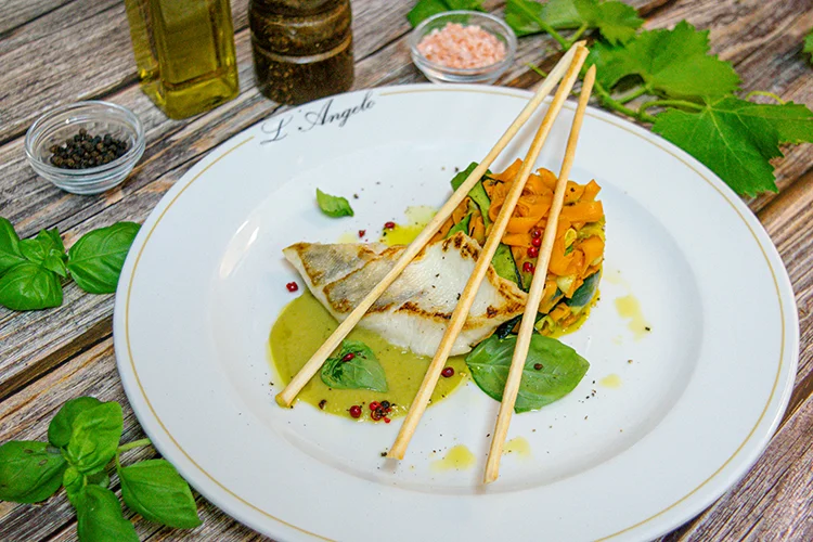 Ein Gourmetgericht auf einem weißen Teller mit gegrilltem Fisch, sautiertem Gemüse, Basilikumblättern und drei Brotstangen, garniert mit Sauce und rosa Pfefferkörnern. Um den Teller herum stehen Kräuter, Öl und Gewürze auf einem Holztisch.
