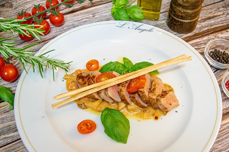 Gekochte Fleischscheiben, serviert auf einem weißen Teller mit einer cremigen Sauce, garniert mit Kirschtomaten, frischem Basilikum und zwei Brotstangen. Kräuter, Kirschtomaten und Gewürze sind um den Teller herum angeordnet.
