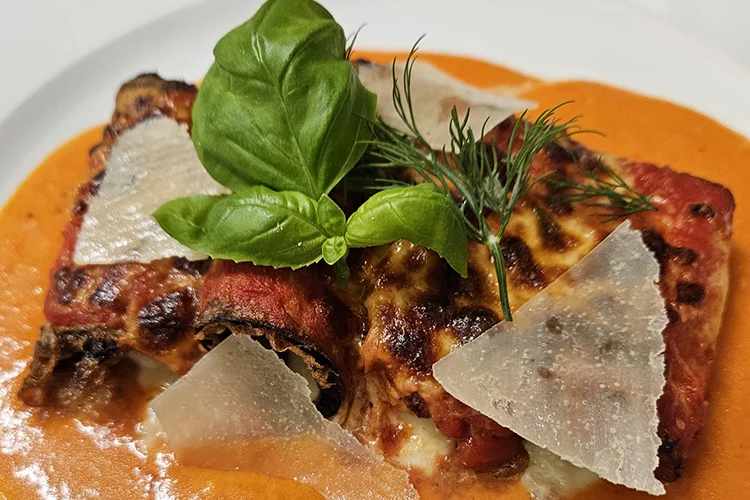Eine Nahaufnahme von gebackenen Auberginenröllchen mit Tomatensauce und geschmolzenem Käse, serviert auf einem Bett aus cremiger Orangensauce, garniert mit frischem Basilikum, Dill und gehobeltem Parmesan.