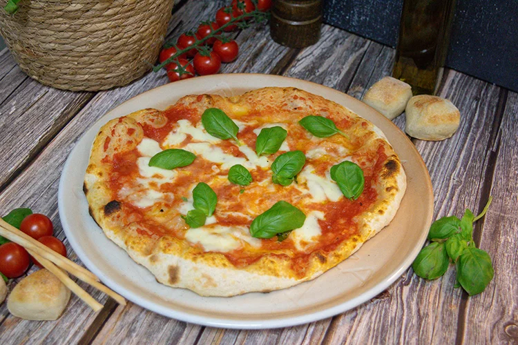 Eine Pizza Margherita mit geschmolzenem Käse, Tomatensauce und frischen Basilikumblättern liegt auf einem Teller. Um sie herum liegen Kirschtomaten, Brotstangen, Basilikum und kleine Brötchen auf einem rustikalen Holztisch.