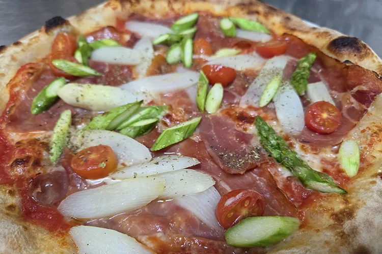 Nahaufnahme einer Pizza mit weißem und grünem Spargel, Kirschtomaten, Prosciutto, Tomatensoße, Käse und etwas schwarzem Pfeffer auf einer goldbraunen Kruste.