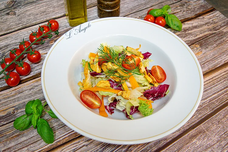 Ein weißer Teller mit einem frischen Salat aus Kopfsalat, Kirschtomaten, Karottenstreifen und Dill auf einem Holztisch, umgeben von Kirschtomaten an der Rebe, Basilikumblättern und Flaschen mit Öl und Pfeffer.