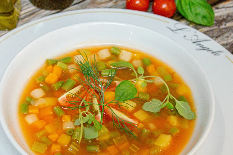 Eine Schüssel Gemüsesuppe mit gewürfelten Karotten, grünen Bohnen und anderem Gemüse, garniert mit frischen Kräutern, serviert auf einem weißen Teller mit der Aufschrift "L'Argile" am Rand. Im Hintergrund sind Tomaten und Basilikum zu sehen.
