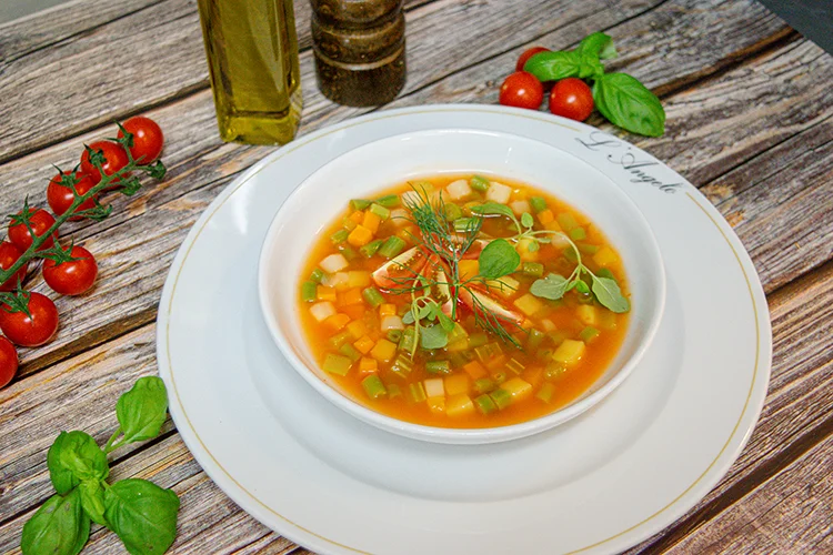 Eine Schüssel Gemüsesuppe mit gewürfelten Karotten, Zucchini und Kräutern steht auf einem weißen Teller, umgeben von frischem Basilikum, Kirschtomaten, Olivenöl und Pfeffer auf einem Holztisch.
