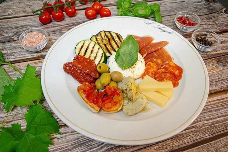 Ein weißer Teller mit verschiedenen italienischen Antipasti: gegrillte Zucchini, Wurstwaren, Käsescheiben, sonnengetrocknete Tomaten, Oliven, Artischocken, Bruschetta und Burrata, garniert mit frischem Basilikum. Zutaten und Kräuter umgeben den Teller.